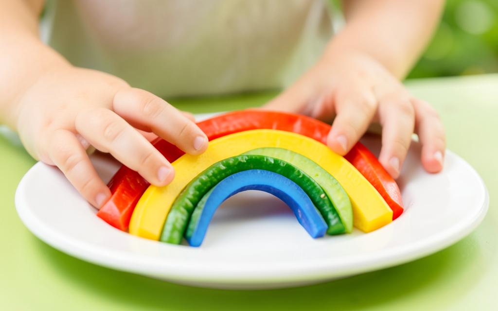 Mãos de criança explorando vegetais coloridos em formato de arco-íris